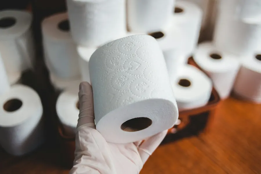 Gloved hand holding a fresh toilet roll in front of stacked rolls